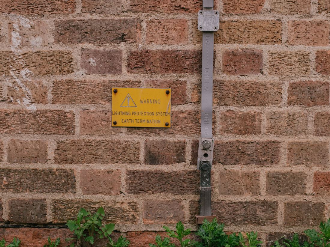 Earth termination strap and a yellow warning sign reading Lightning Protection System Earth Termination on a brick wall