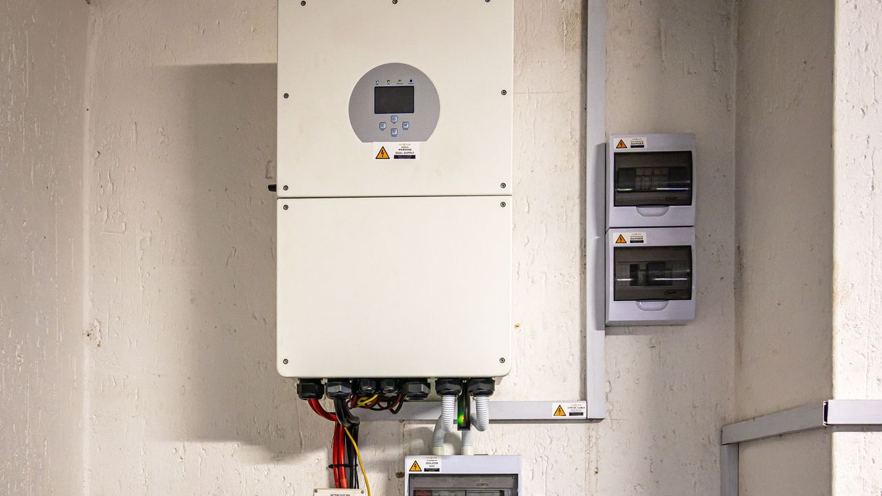 An off-grid solar installation: a wall-mounted hybrid inverter with a built-in MPPT charge controller, isolators, and a fuse, sitting above a stacked lithium battery bank with red and black cabling