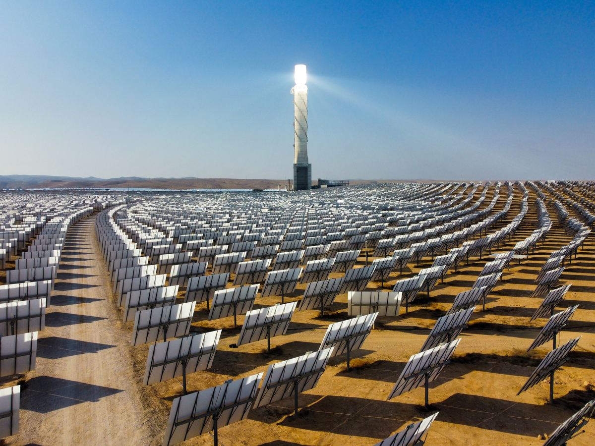 Aerial view of a concentrated-solar-power plant in the desert: thousands of mirrors arranged in curved rows around a brightly lit central receiver tower under a clear blue sky