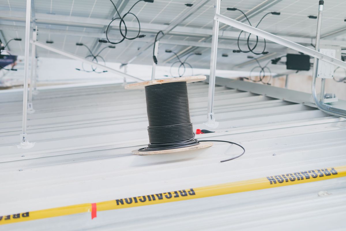 A spool of black DC solar cable on a metal rooftop beneath a solar panel array, with panel wiring looped above