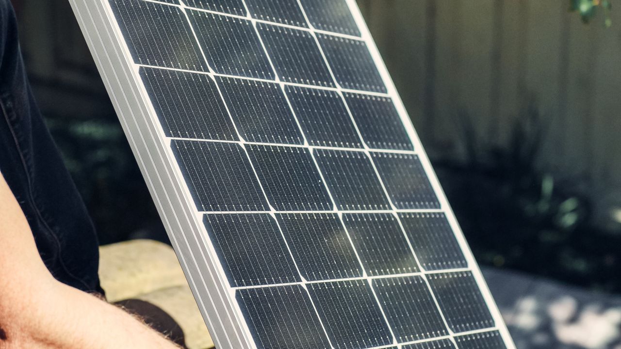 A person in work gloves holding a portable 100W monocrystalline solar panel outdoors in bright sun